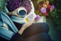 Person holding a cup of coffee and a smartphone, surrounded by flowers outdoors.